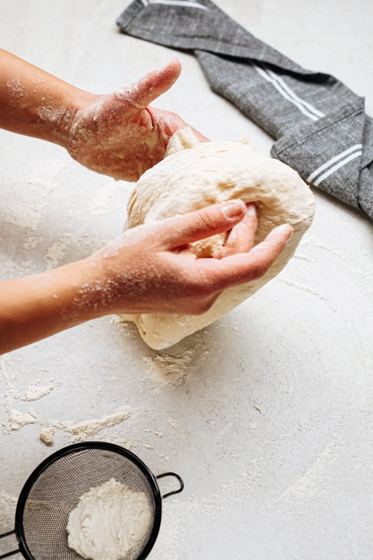 Two hands kneading a ball of dough on a floured surface, with a gray-striped kitchen towel and a small mesh strainer in the background.