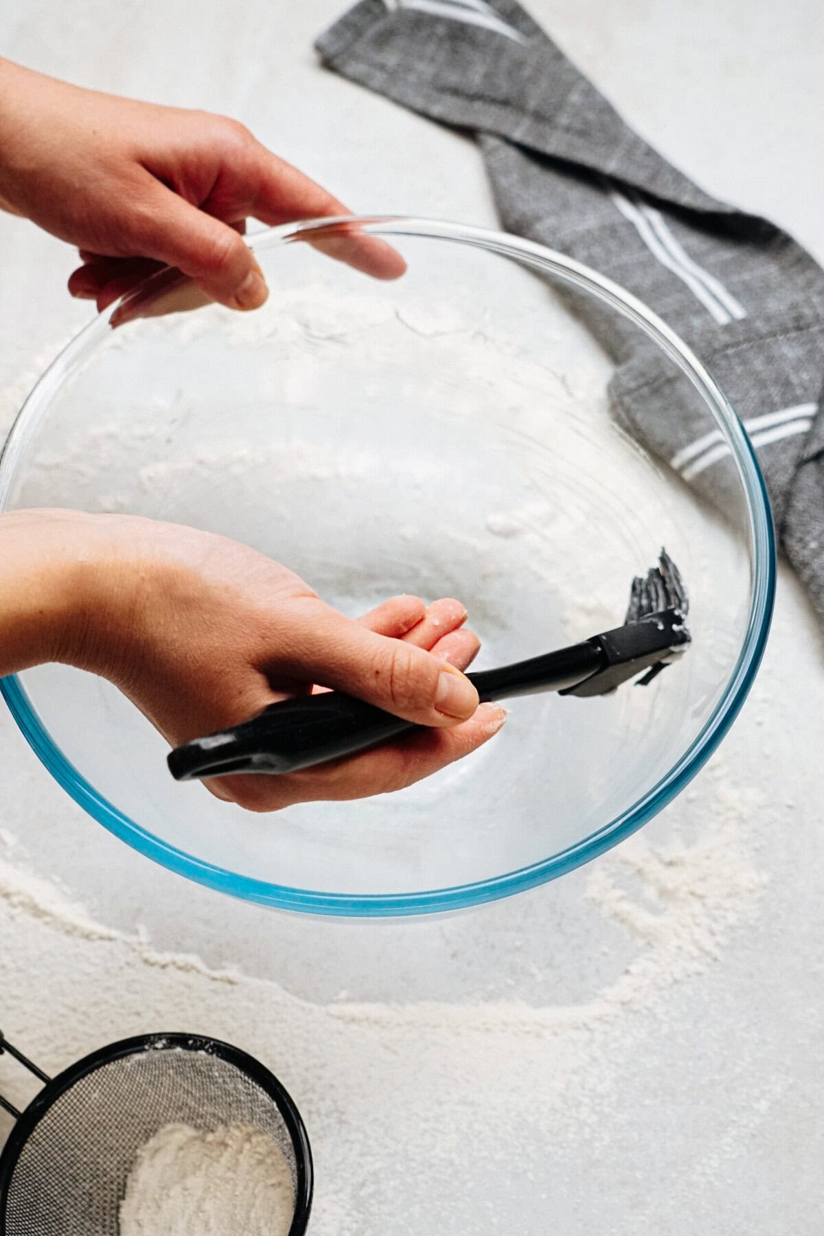 Hands coating a glass mixing bowl with oil using a brush. Flour and a sift are visible on the countertop.