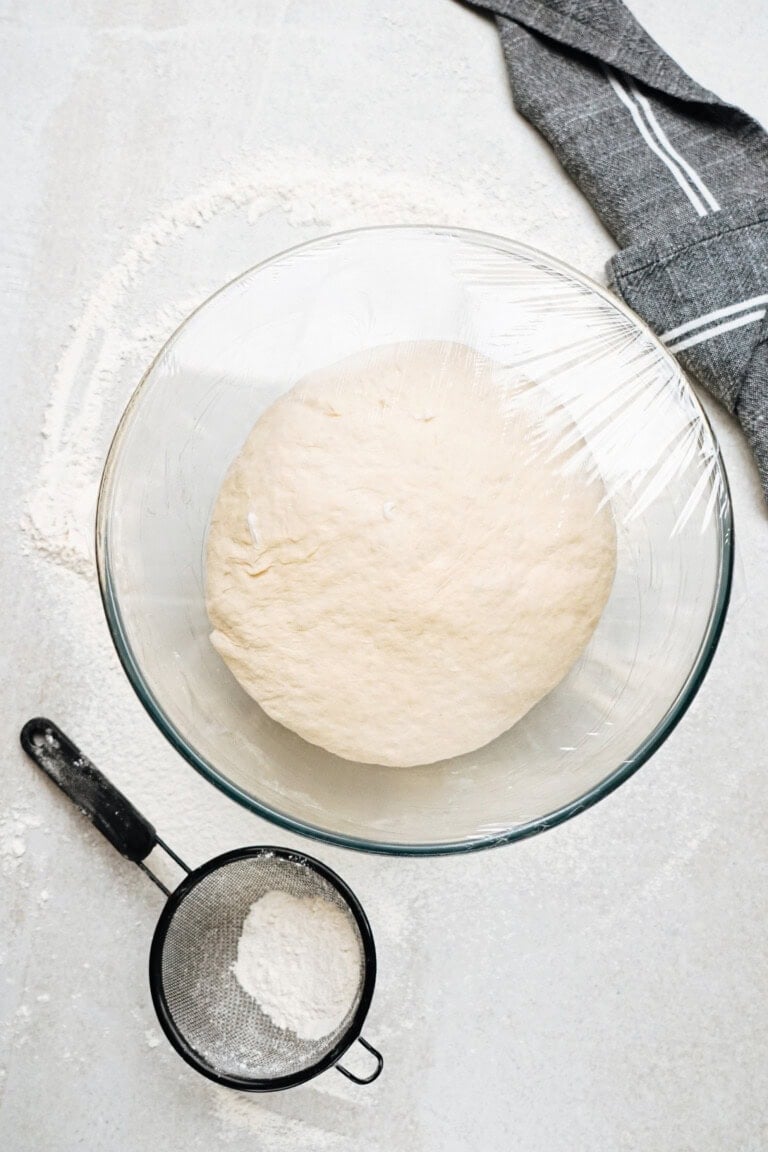 A bowl of dough covered with plastic wrap is on a floured surface next to a sifter and a gray cloth.