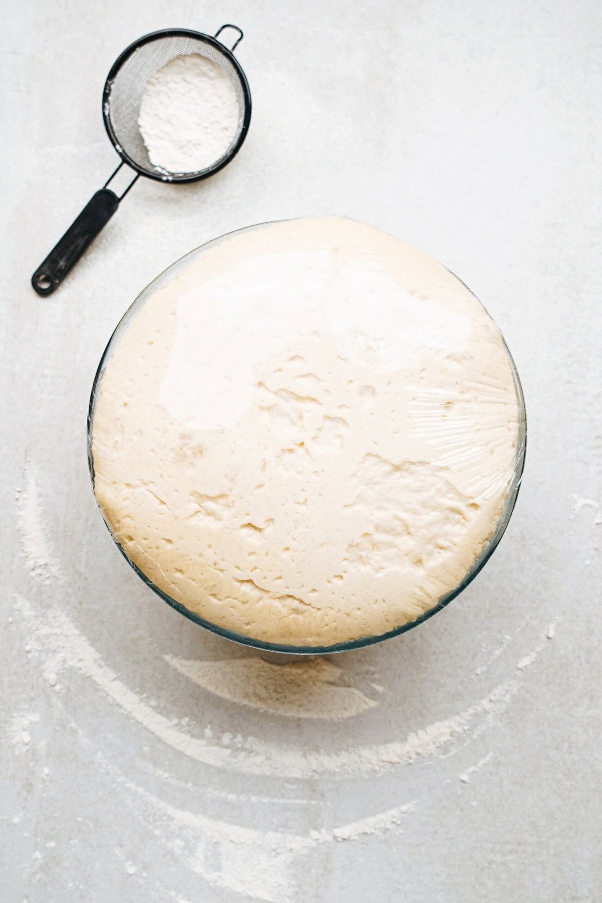 A bowl of risen dough covered in plastic wrap with a small sifter filled with flour beside it on a light-colored surface.