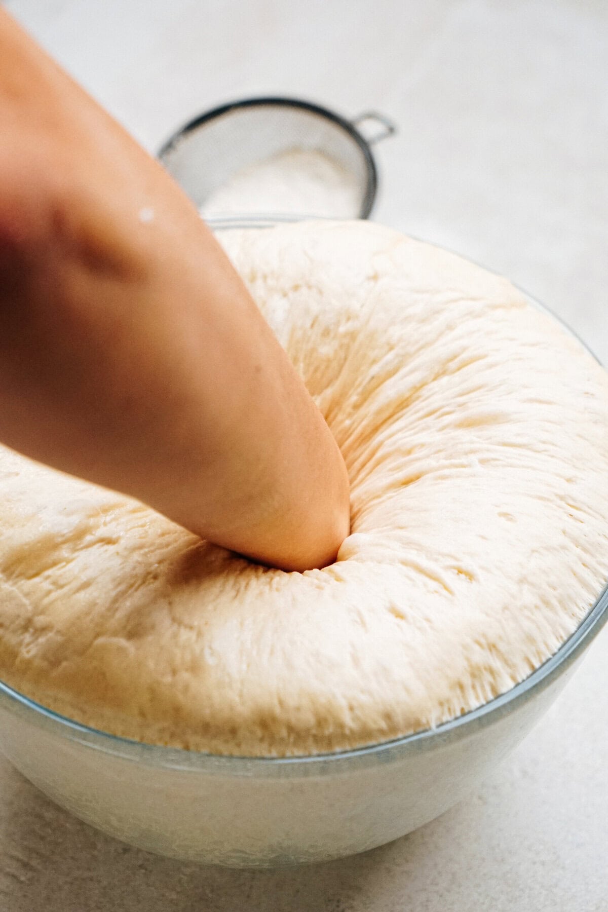 A hand is seen punching down a large, risen ball of dough in a clear glass bowl on a light-colored surface. A sifter is visible in the background.