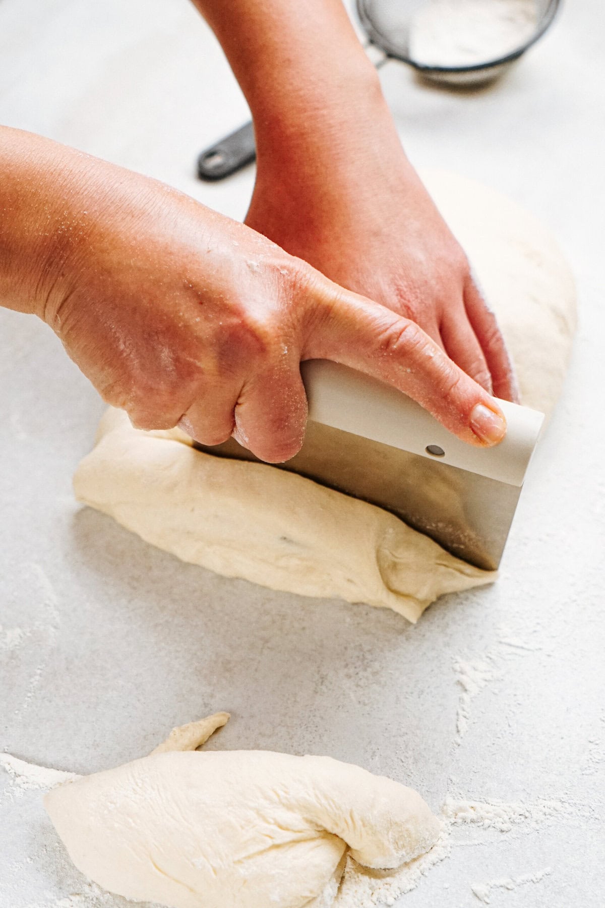 A pair of hands use a bench scraper to cut a portion of dough on a floured surface, with additional flour and dough pieces visible in the background.