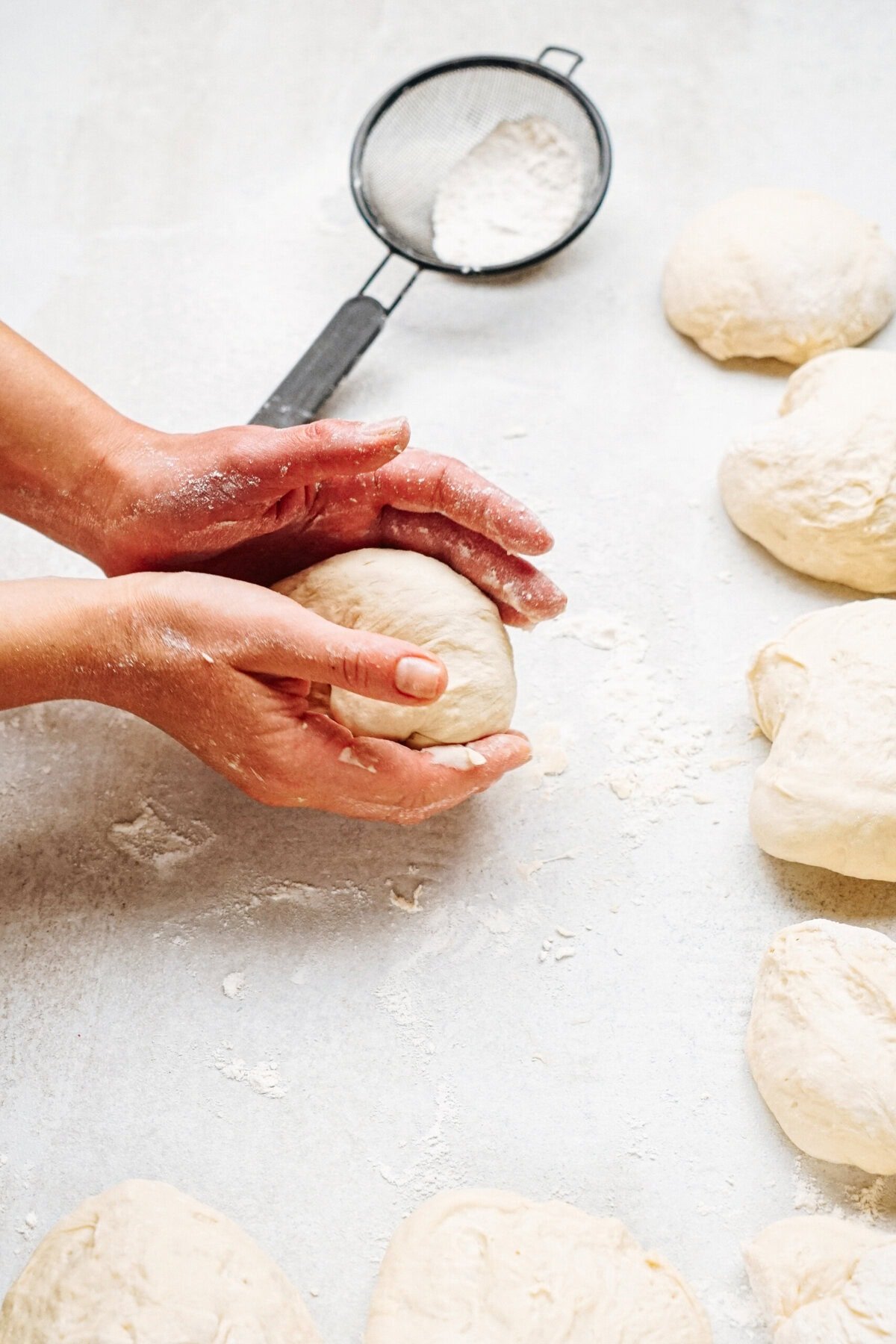 Hands shaping dough on a floured surface with several dough balls and a flour sifter nearby.