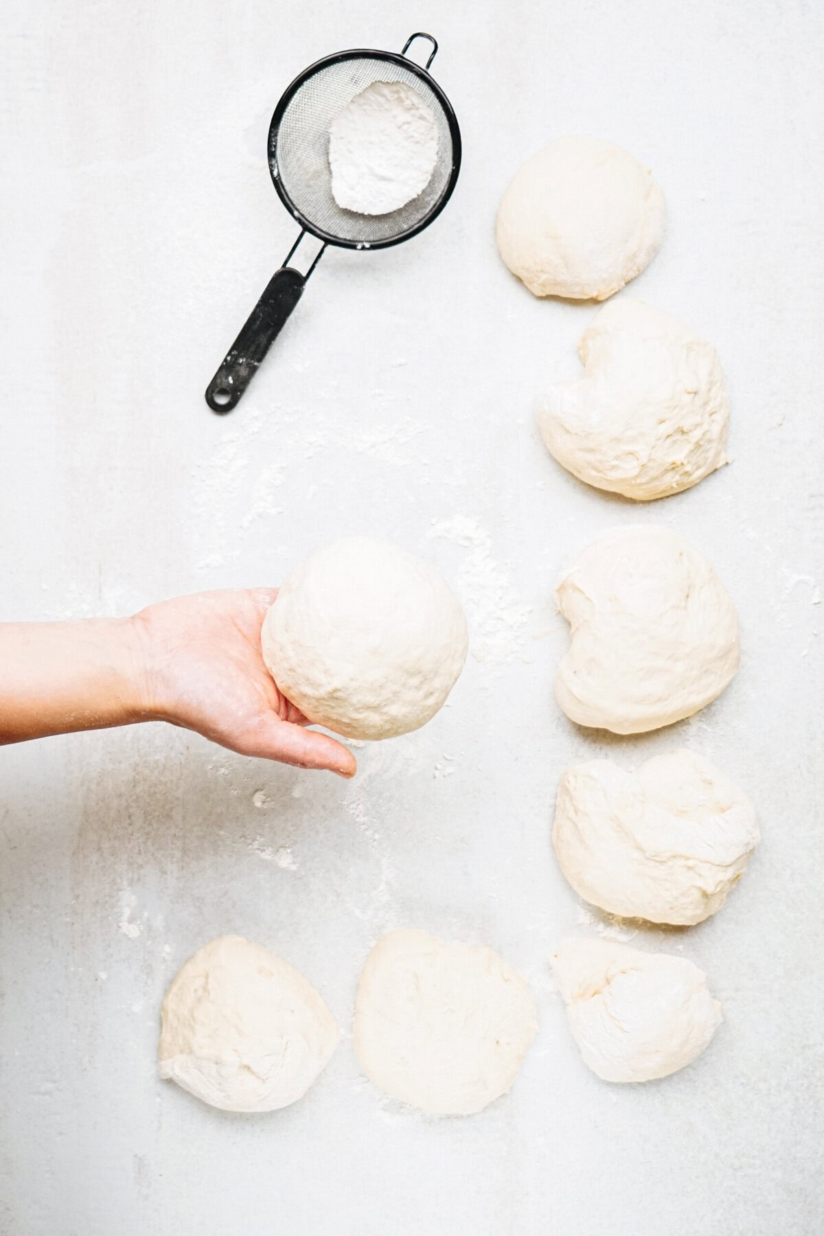 A hand holding a ball of dough, with additional dough pieces and a flour sifter on a white surface.