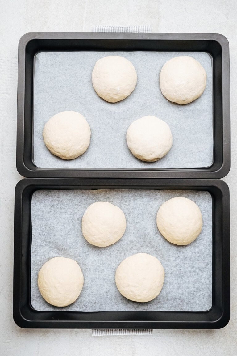 Two baking trays each hold three round dough balls placed on parchment paper, arranged in a 2x3 grid pattern.