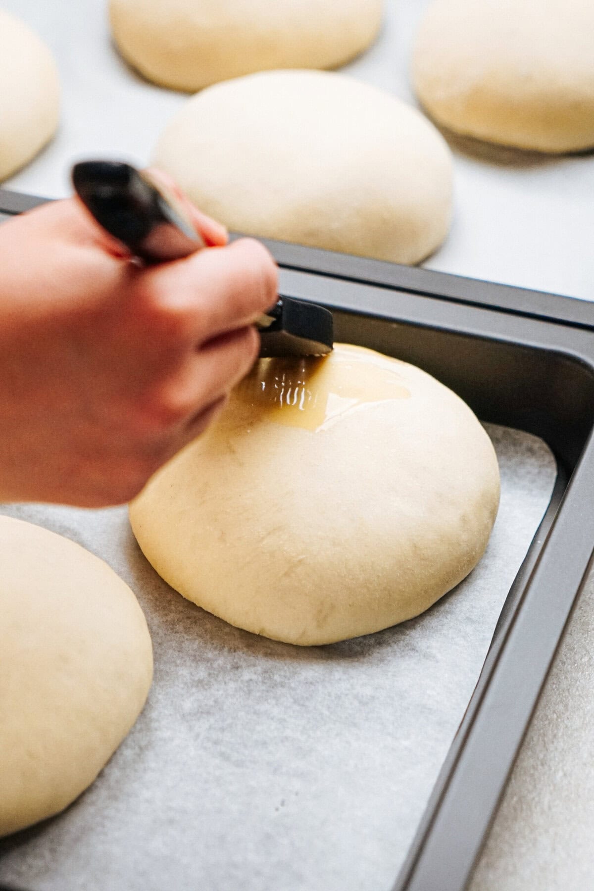 A hand is brushing egg wash on a round dough ball on a parchment-lined baking tray, with more dough balls in the background.