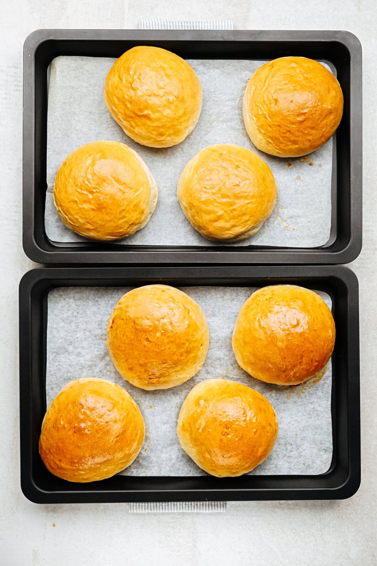 Two black baking trays each containing four golden brown buns on parchment paper, arranged on a white surface.