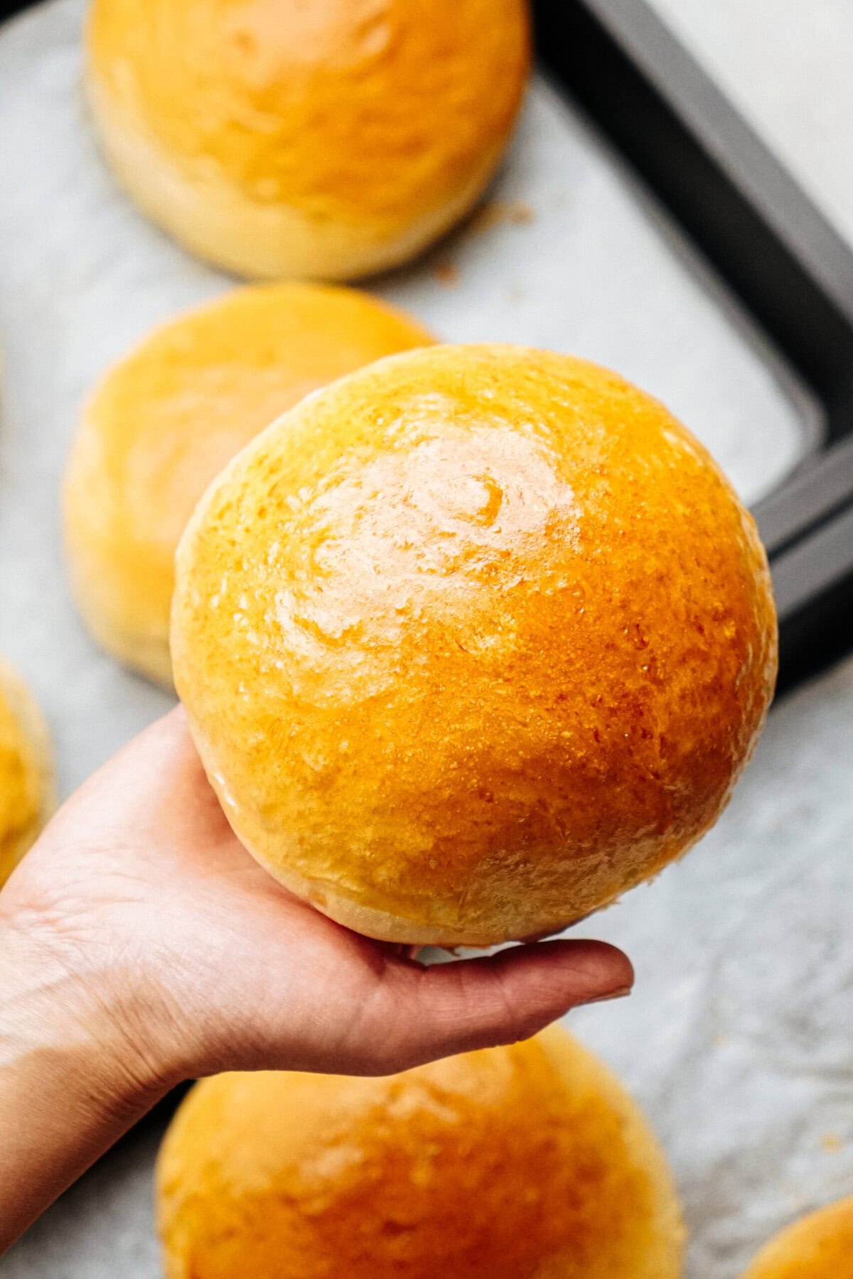 A hand holding a freshly baked, golden brown bun with more buns on a baking tray in the background.