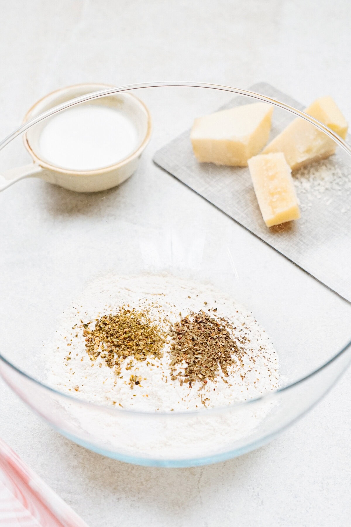 A clear glass bowl contains flour with two piles of ground pepper or spices on top. Nearby, a small bowl holds a white substance, and several blocks of cheese are on a cutting board.