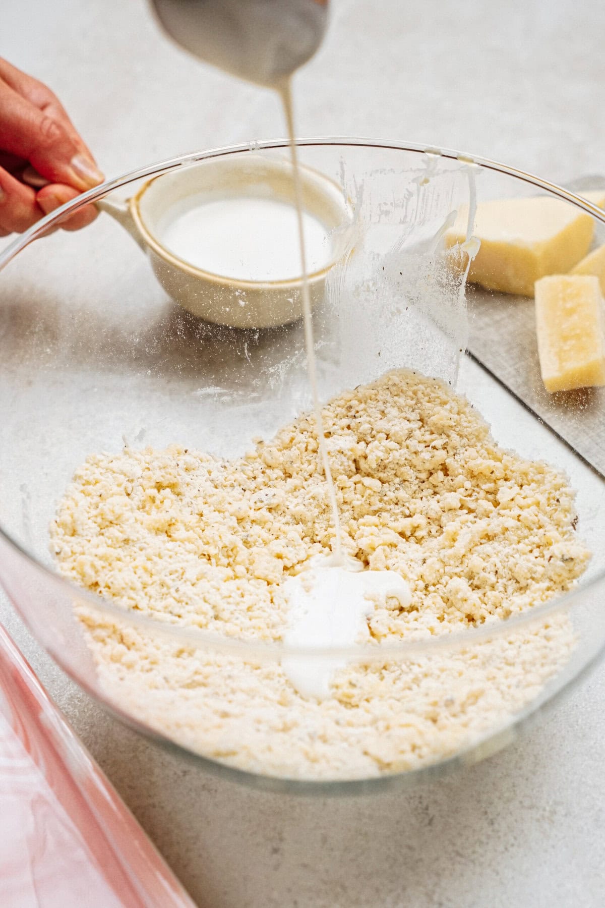 A hand pours liquid from a cup into a large glass mixing bowl containing a crumbly mixture, with another cup of liquid and a block of cheese visible in the background.