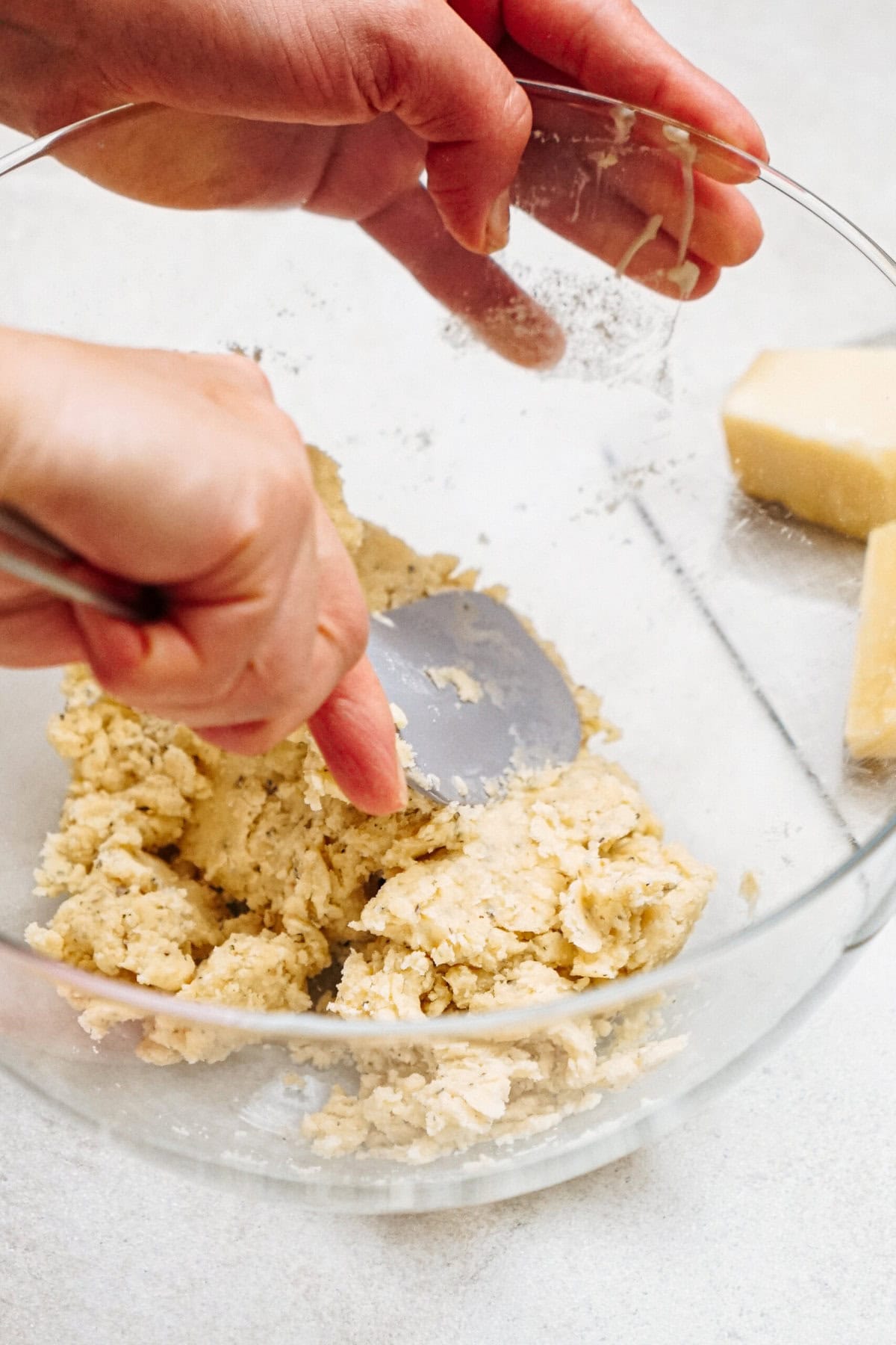 Hands mixing dough in a clear glass bowl with a utensil, with blocks of cheese visible in the background.