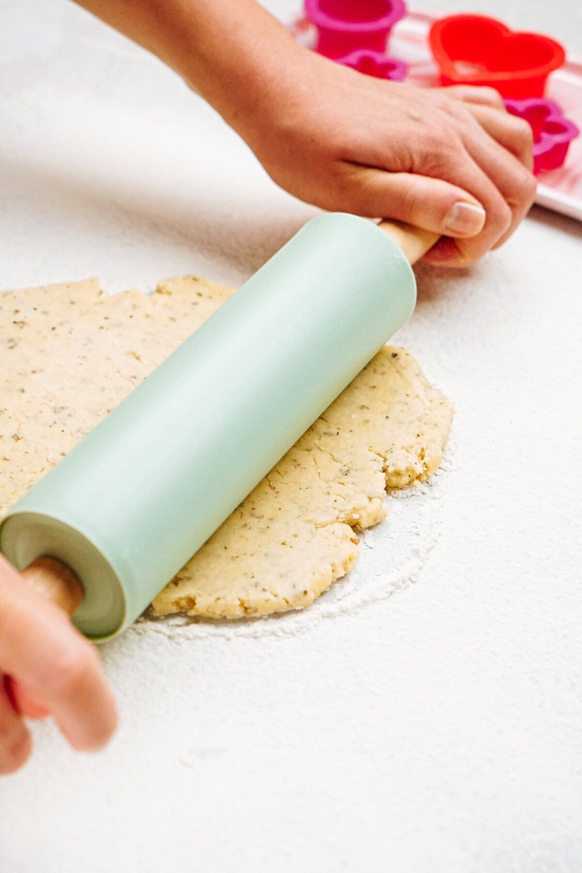 Hands rolling out cookie dough with a green rolling pin; heart-shaped cookie cutters are visible in the background.