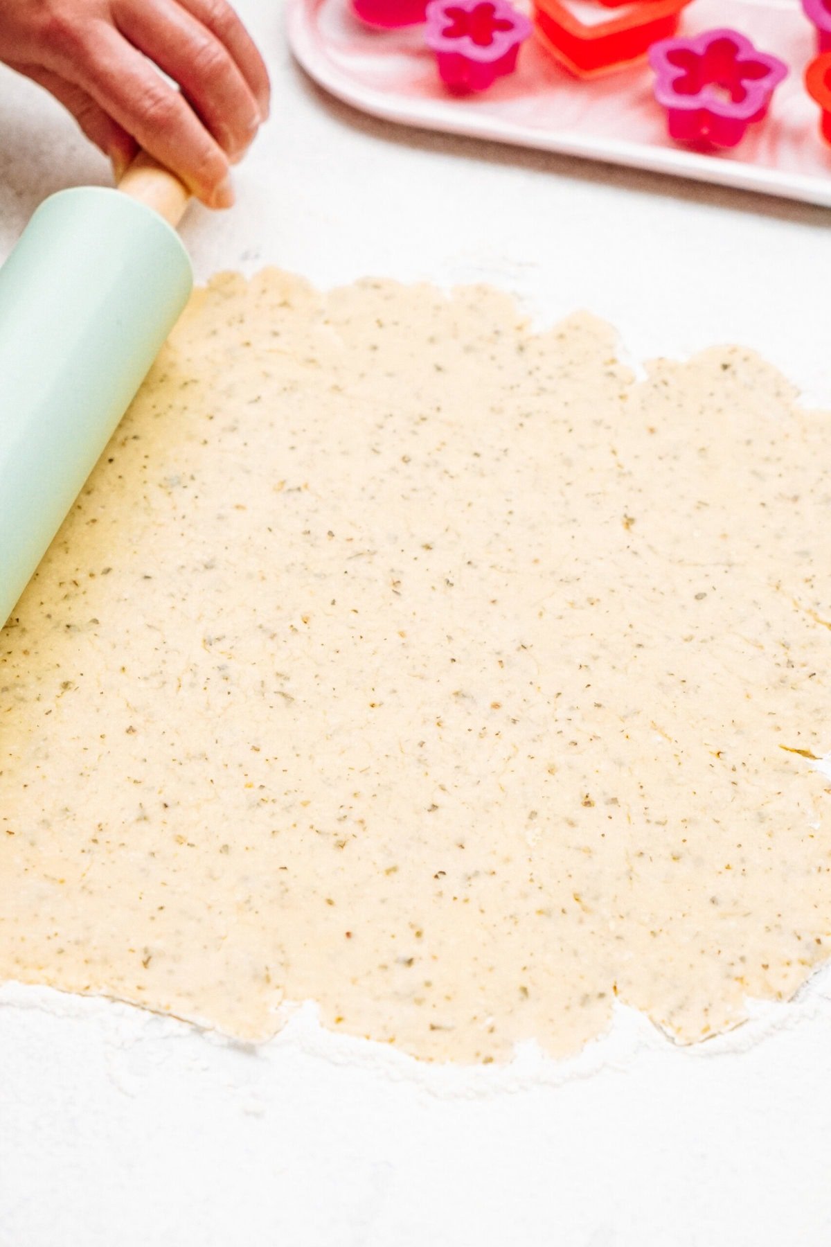 A hand rolling out cookie dough with a rolling pin on a counter. Flower-shaped cookie cutters are visible in the background.