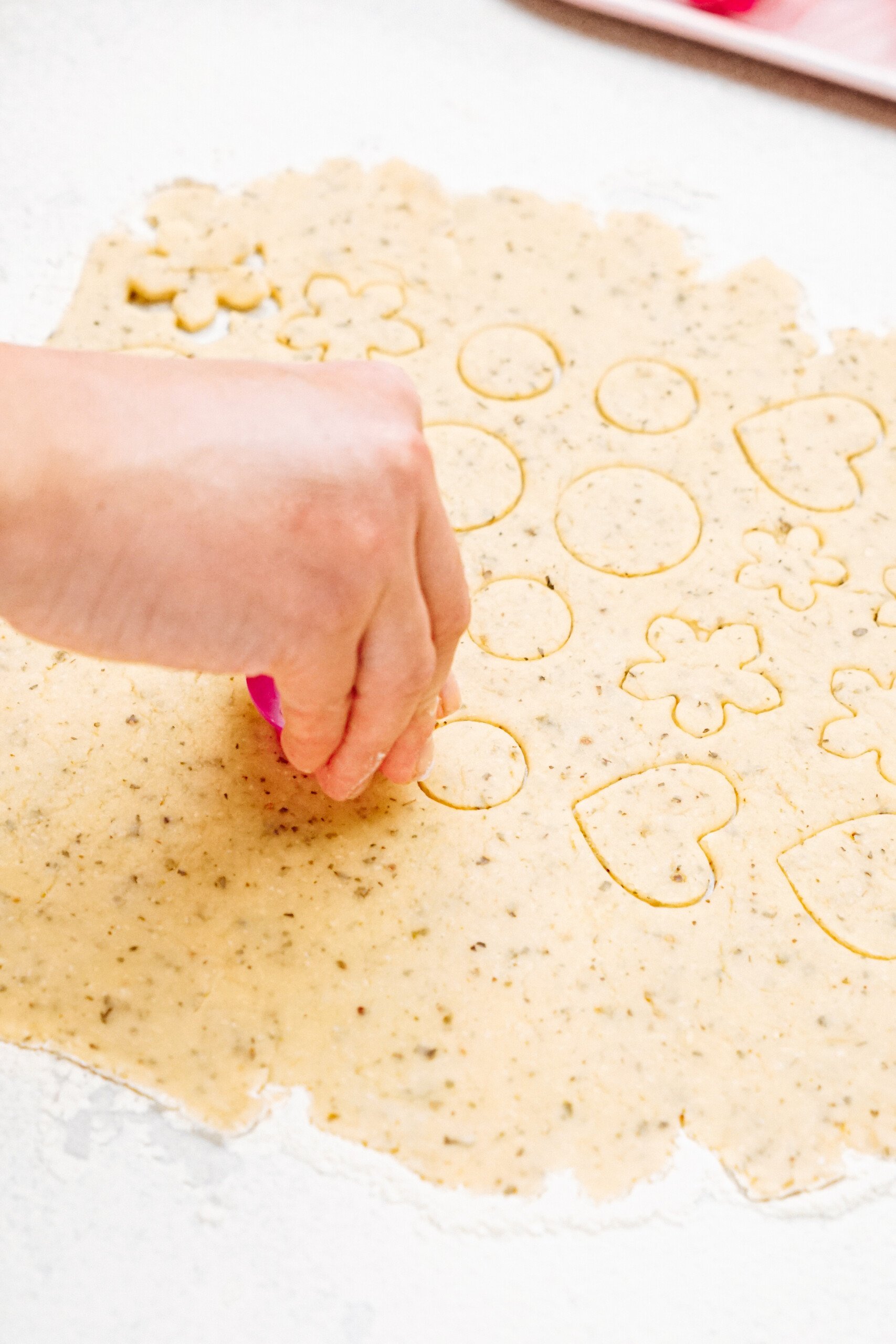 A hand uses a cookie cutter to shape dough on a flat surface, creating various shapes including circles, hearts, and flowers.