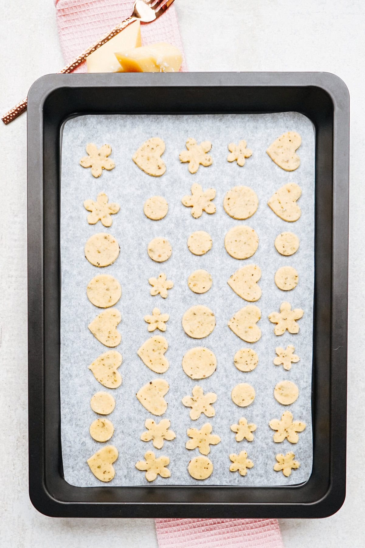 A baking tray lined with parchment paper holds an assortment of small, raw dough shapes including flowers, circles, stars, and semi-circles arranged in neat rows.