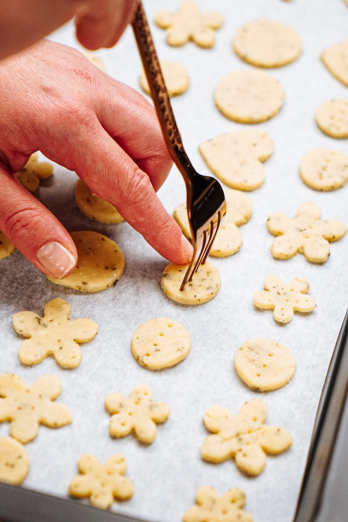 A hand uses a fork to prick small round and flower-shaped dough pieces placed on a parchment-lined baking sheet.