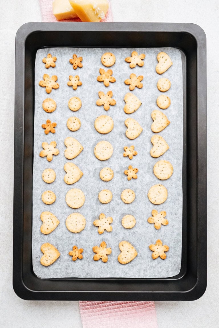 A baking tray lined with parchment paper holds an assortment of small, baked crackers in various shapes including hearts, flowers, stars, and circles.