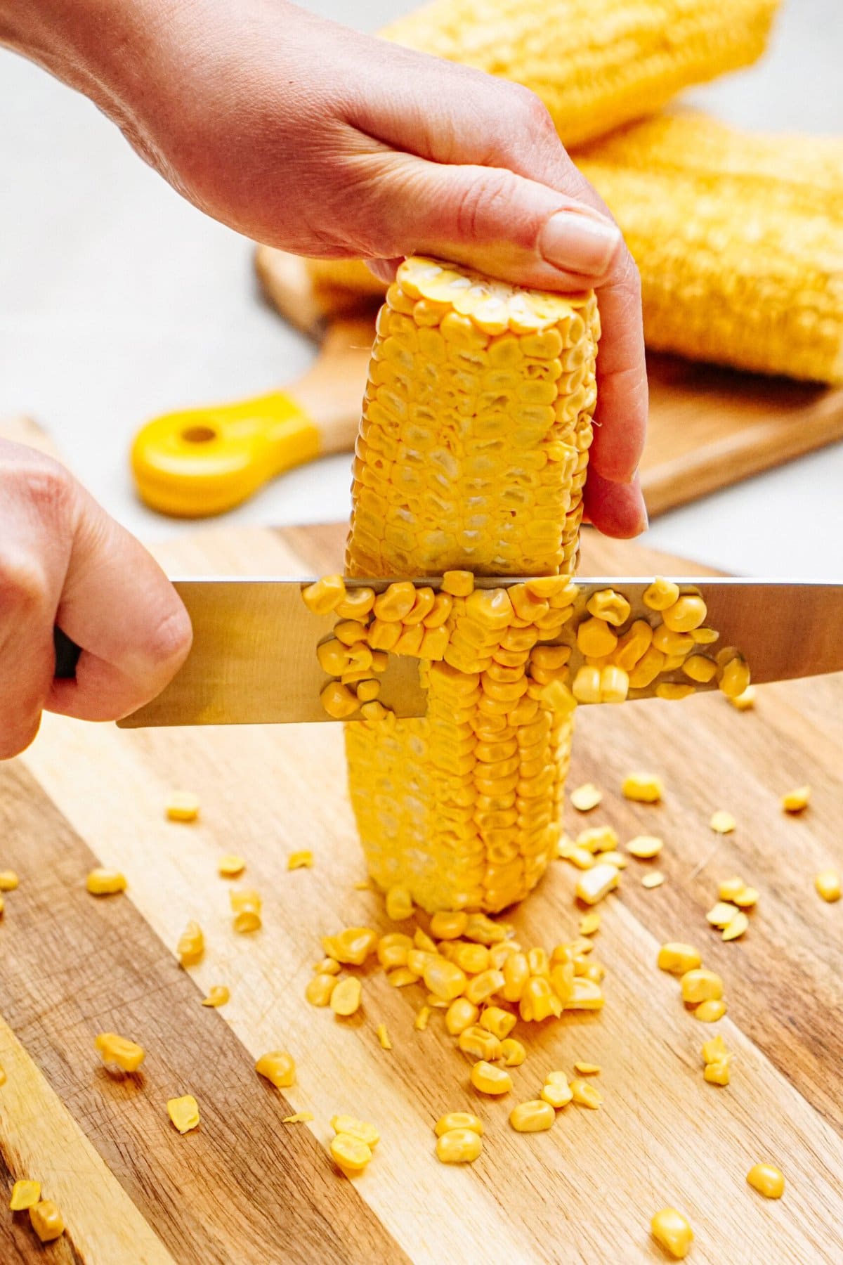 A person is using a knife to cut corn kernels off a cob on a wooden cutting board, with another cob in the background, preparing fresh ingredients for chicken corn chowder.