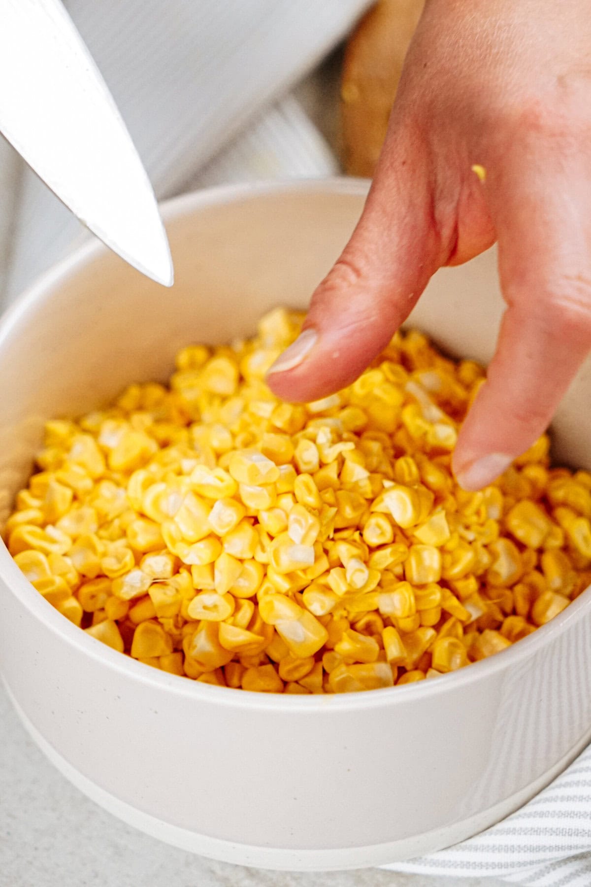 A hand reaching into a bowl filled with corn kernels, with a knife nearby, as if preparing to make chicken corn chowder.