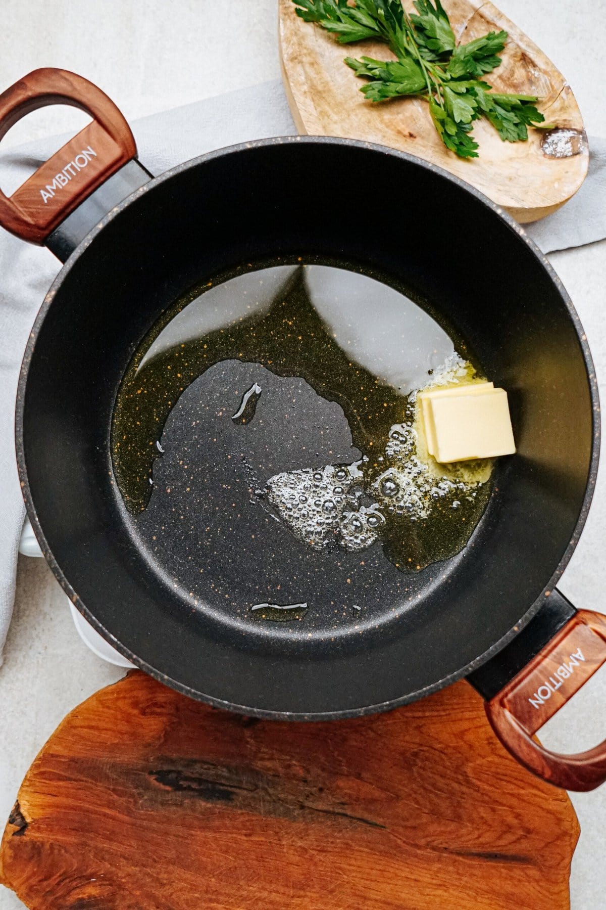 A black pot with "Ambition" on its handles sits on a wooden surface. Inside, a pat of butter is melting in oil, the start of a hearty chicken corn chowder. A wooden cutting board with parsley is partially visible in the background.