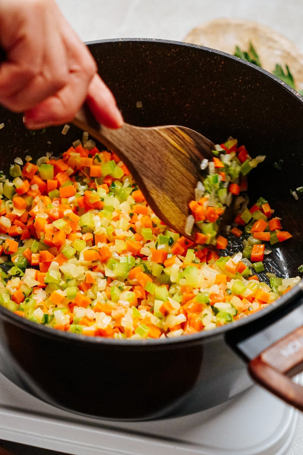 A person stirs chopped vegetables, including carrots, celery, and onions, in a black cooking pot with a wooden spoon, preparing the hearty chicken corn chowder.