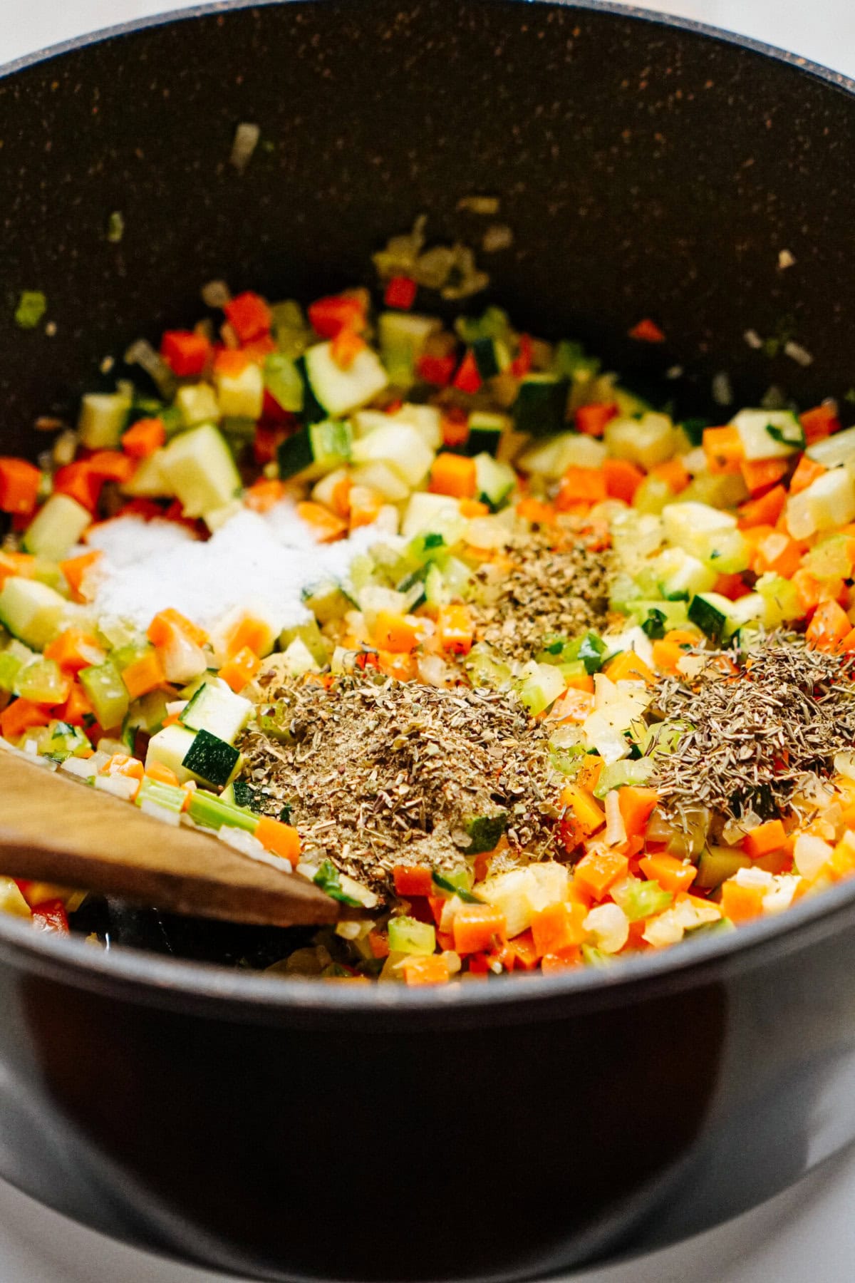 Diced vegetables and various spices are cooking in a black pot, blending into a savory chicken corn chowder as a wooden spoon stirs the mixture.