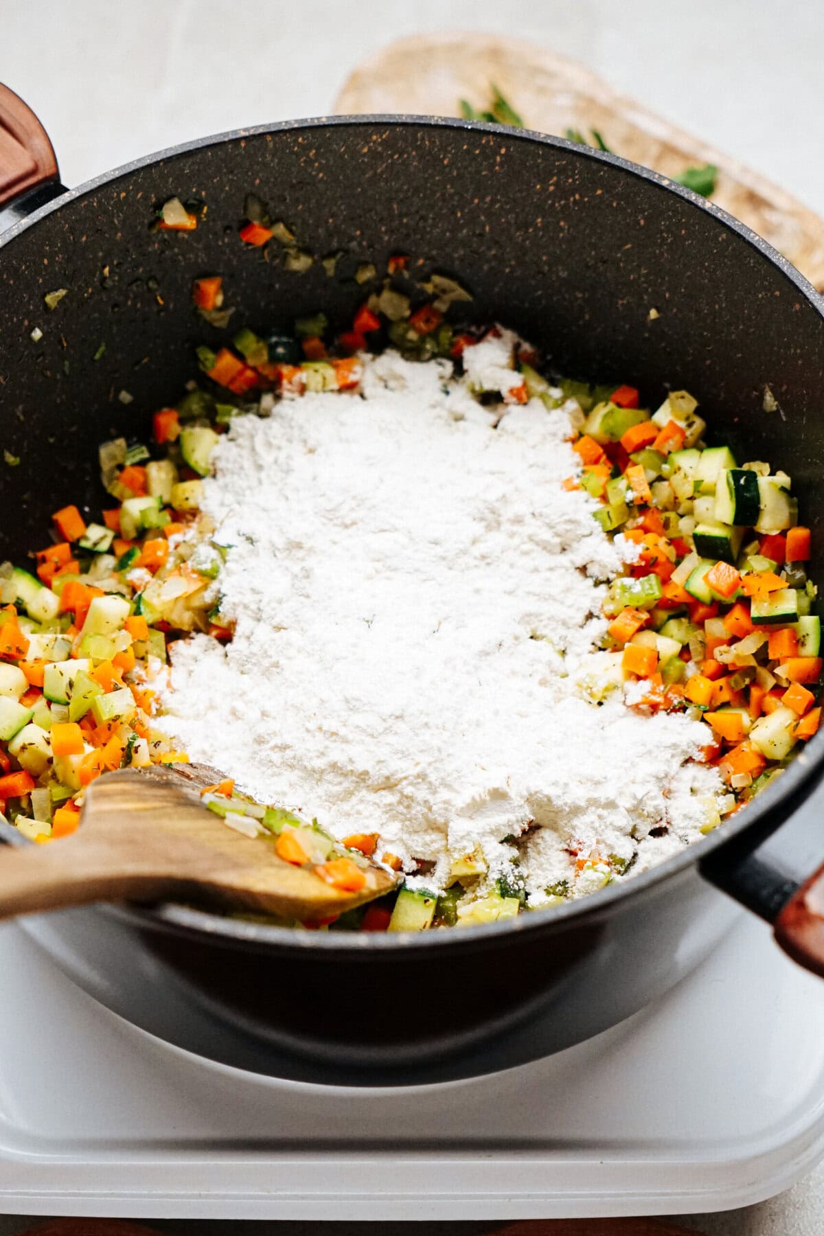 A pot containing a mixture of diced vegetables and flour, reminiscent of a hearty chicken corn chowder, with a wooden spatula resting on the rim.