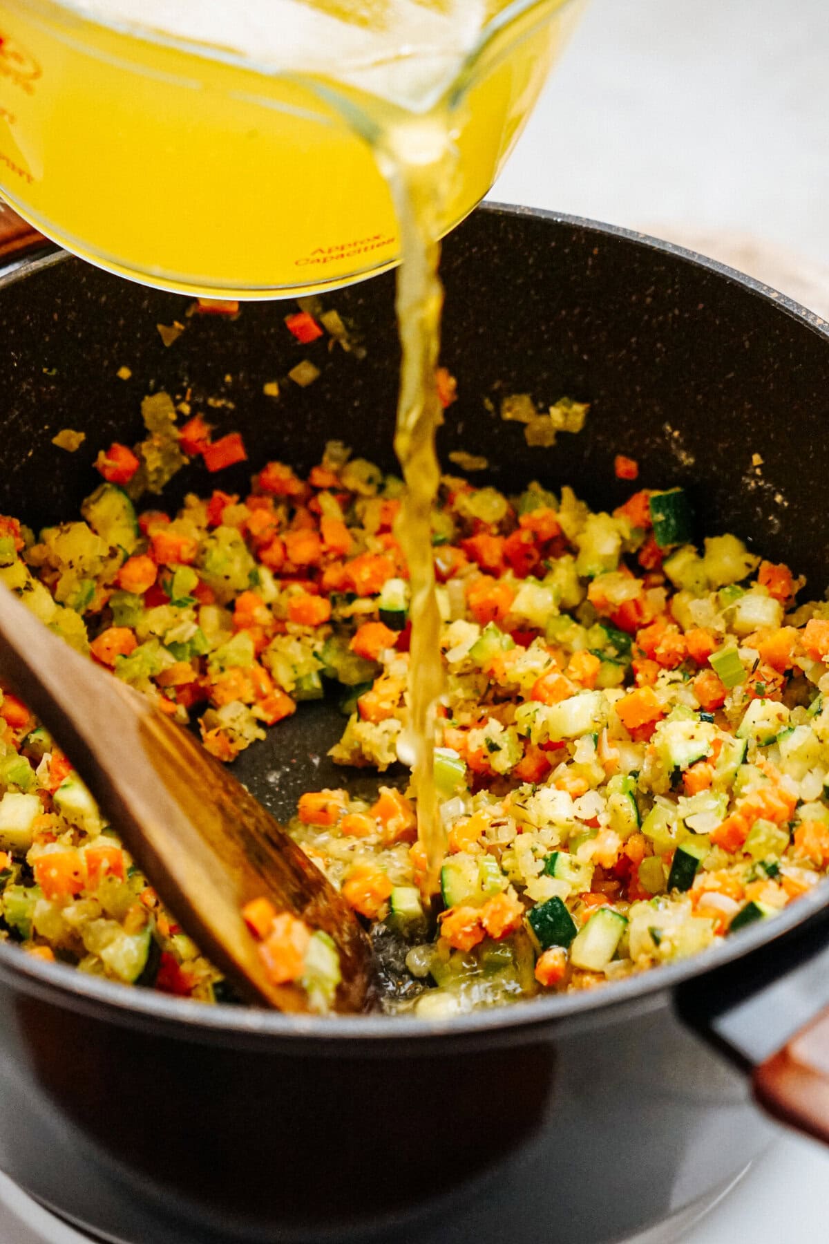 A pot on the stove containing chopped vegetables being stirred with a wooden spatula as broth is poured in from a measuring cup, creating the perfect base for a hearty chicken corn chowder.