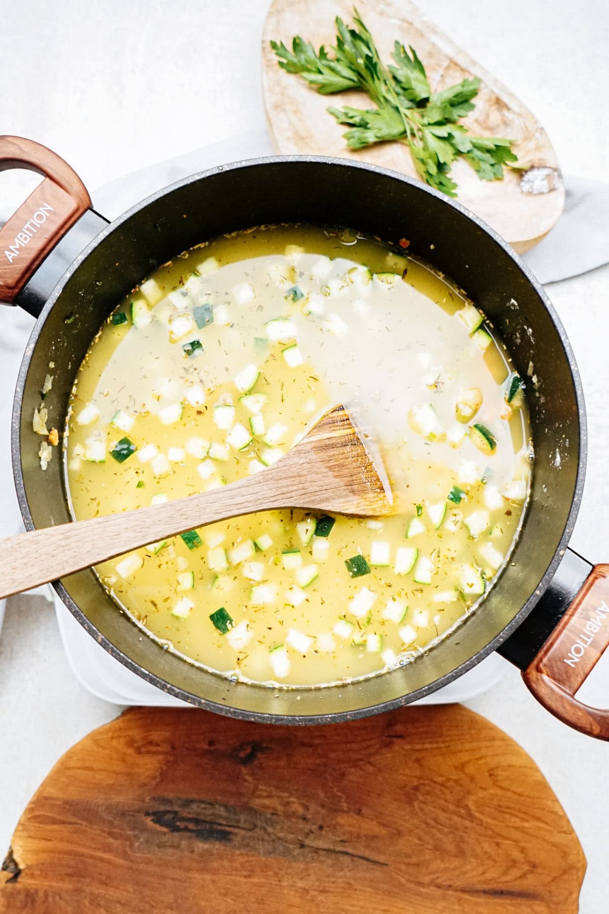 A pot of chicken corn chowder with diced vegetables and a wooden spoon, placed on a wooden cutting board. Fresh herbs are on a plate in the background.