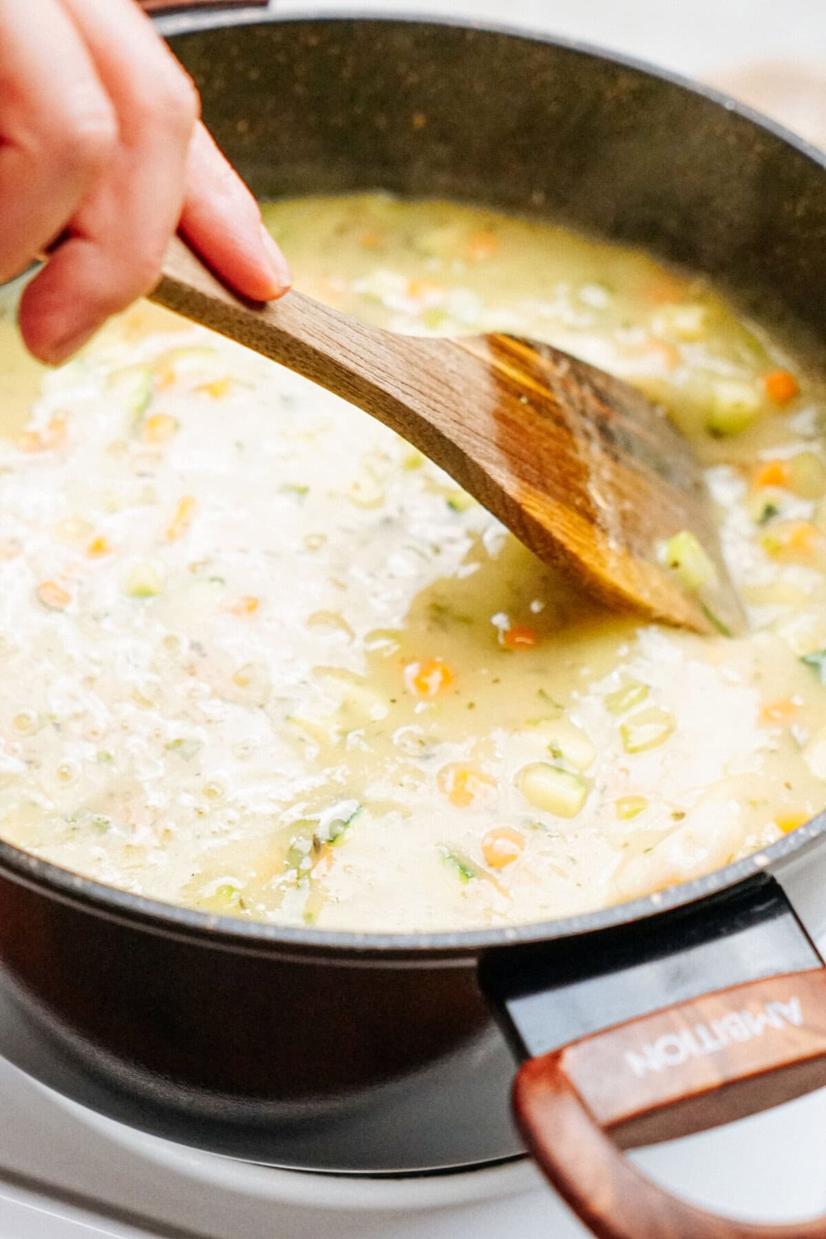 A person stirring a pot of creamy chicken corn chowder with a wooden spoon on a stovetop.