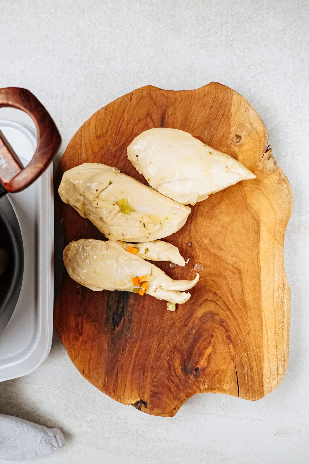 Three cooked chicken breasts rest on a wooden cutting board, with part of a gray pot and a wooden spoon handle visible on the left side of the image, hinting at a hearty chicken corn chowder in progress.