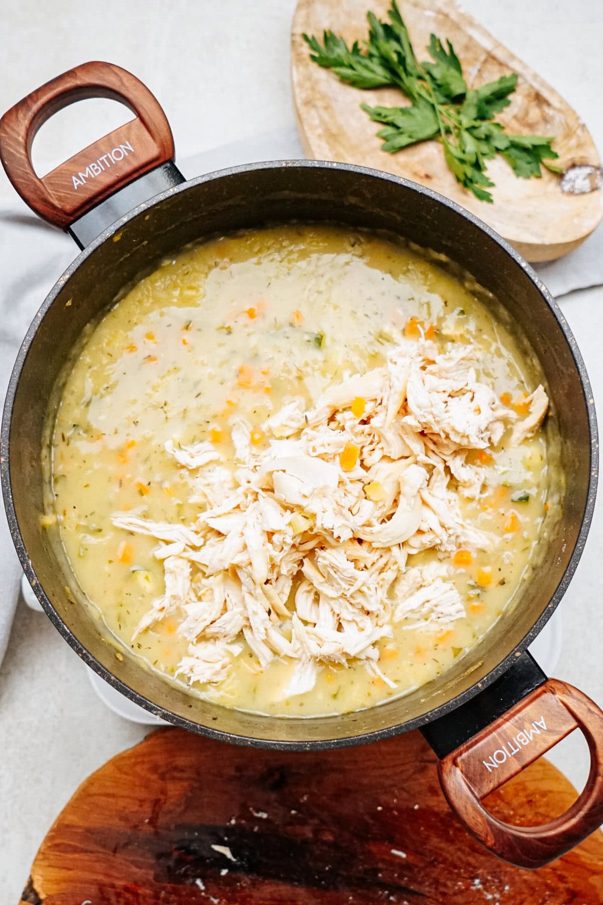 A pot filled with a creamy chicken and vegetable soup, reminiscent of a hearty chicken corn chowder, is placed on a wooden board. Shredded chicken is on top. A wooden board with parsley is in the background.