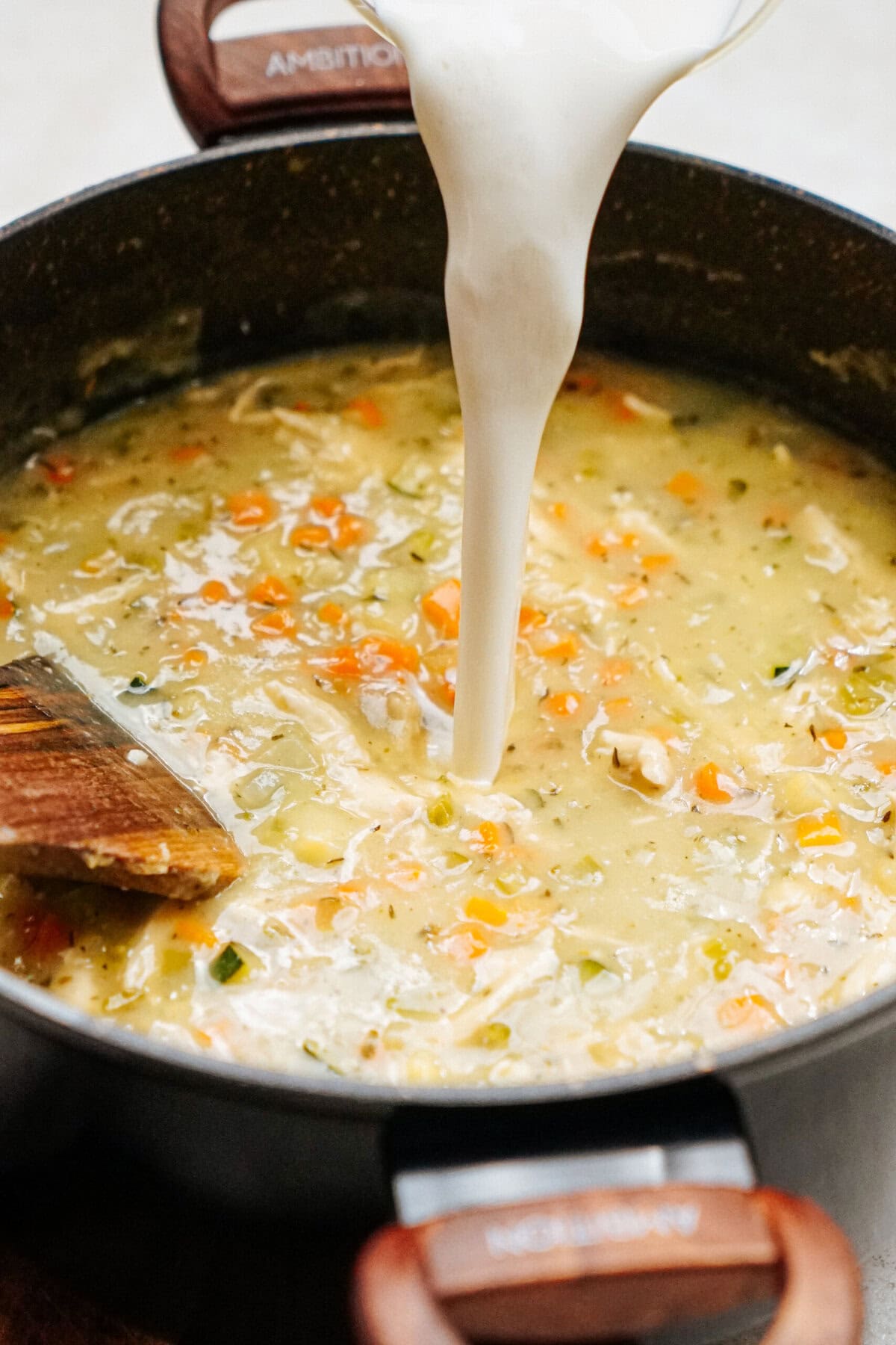 A creamy liquid is being poured into a pot containing a mixture of diced vegetables and broth, creating the perfect base for a delicious chicken corn chowder, being stirred with a wooden spatula.