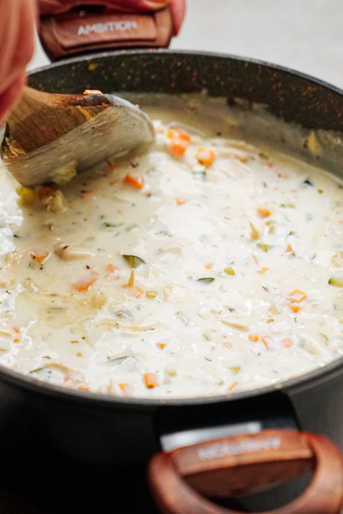 A close-up view of a pot filled with creamy chicken corn chowder, being stirred with a wooden spoon. The pot has wooden handles labeled "AMBITION.