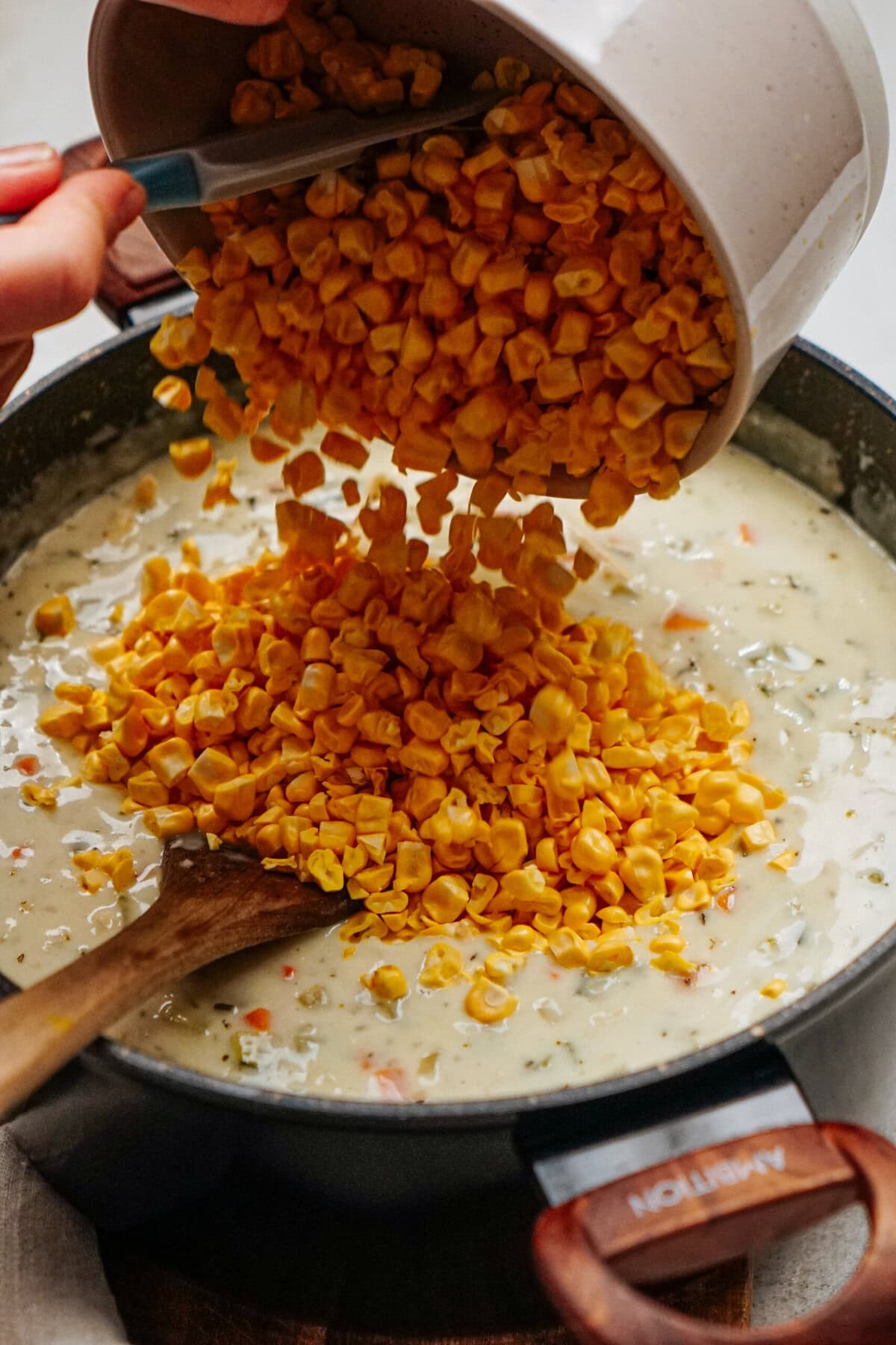 A person is pouring a bowl of corn into a pot of creamy chicken corn chowder being stirred with a wooden spoon.