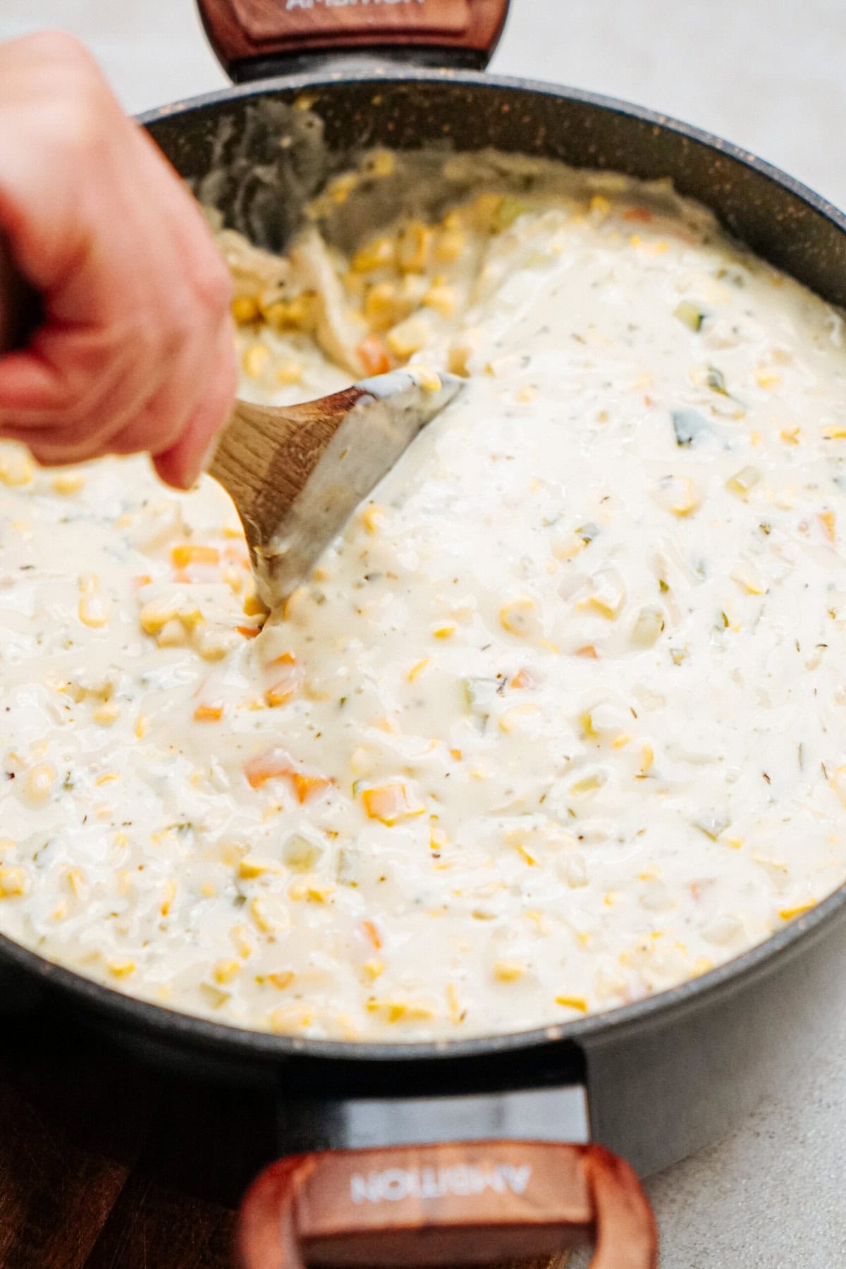 Close-up of a person stirring a creamy chicken corn chowder in a black pot using a wooden spoon. The chowder contains visible corn kernels and chunks of vegetables.