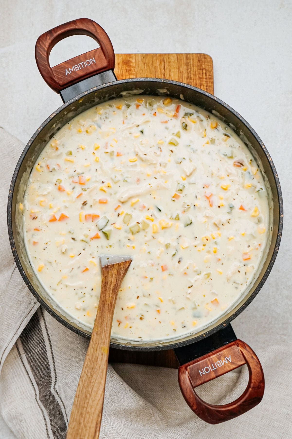 A large pot filled with creamy chicken corn chowder, including carrots and celery. A wooden spoon rests on the rim, and the pot is placed on a wooden cutting board with a cloth underneath.