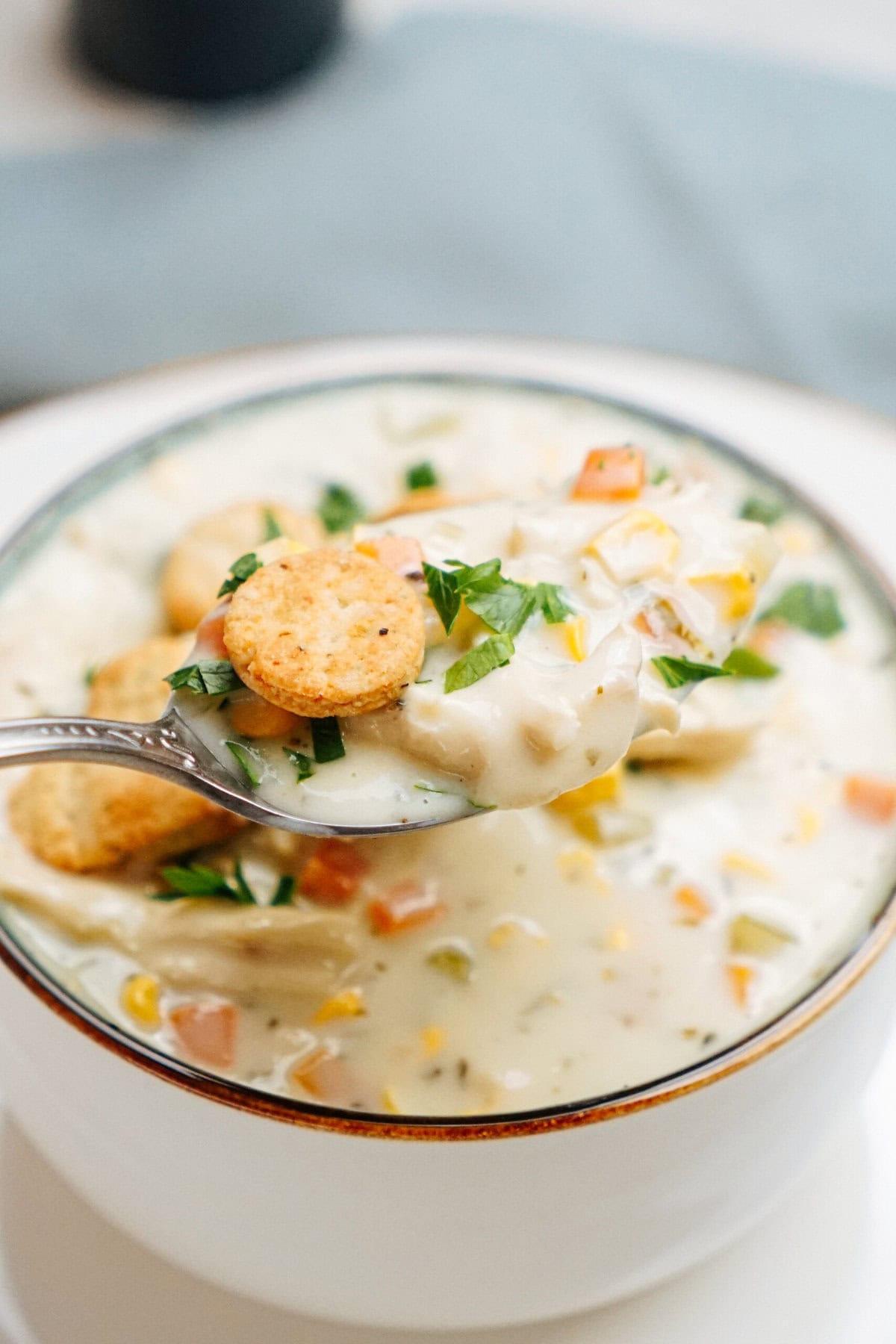 A spoonful of creamy chicken corn chowder with crackers, held above a bowl filled with the same soup, garnished with chopped herbs.