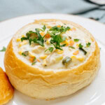 A bread bowl filled with creamy chicken corn chowder, topped with chopped vegetables and herbs, is placed on a white plate. A piece of bread and a spoon are nearby.