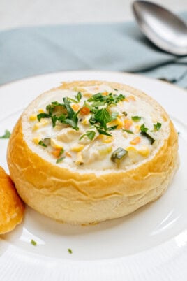 A bread bowl filled with creamy chicken corn chowder, topped with chopped vegetables and herbs, is placed on a white plate. A piece of bread and a spoon are nearby.