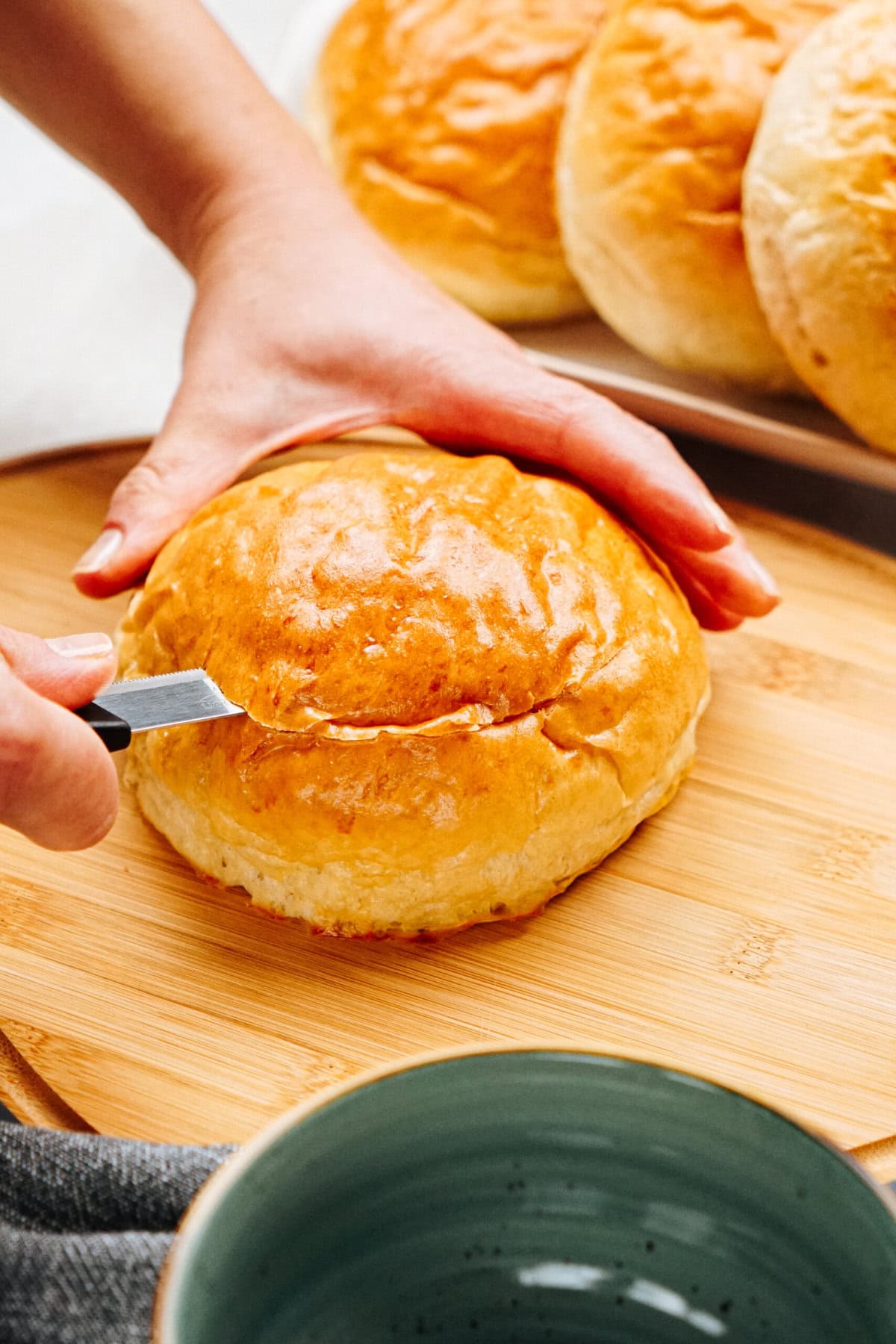 A person uses a knife to slice a round bread roll on a wooden cutting board. Several bread rolls and a green bowl are visible nearby.