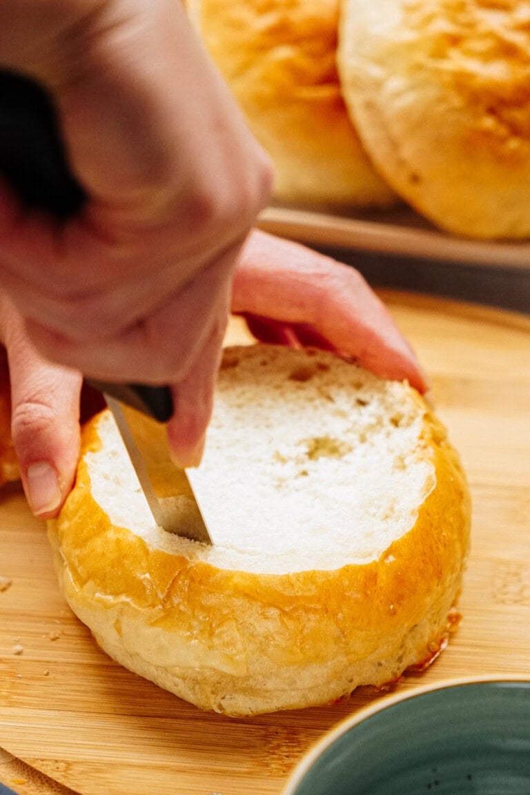 A hand uses a knife to cut into the top of a round bread loaf on a wooden cutting board, with more bread loaves in the background.