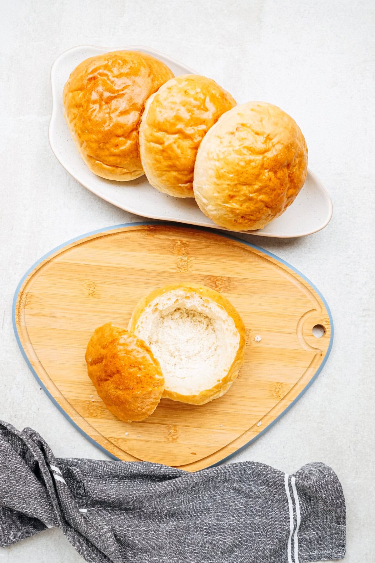 Three round bread rolls on a white ceramic dish, with one bread roll hollowed out on a wooden cutting board beside a cloth.