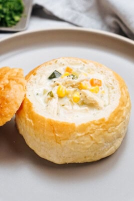 A bowl of creamy soup with vegetables and chicken served in a hollowed-out bread bowl, with the bread top placed on the side.