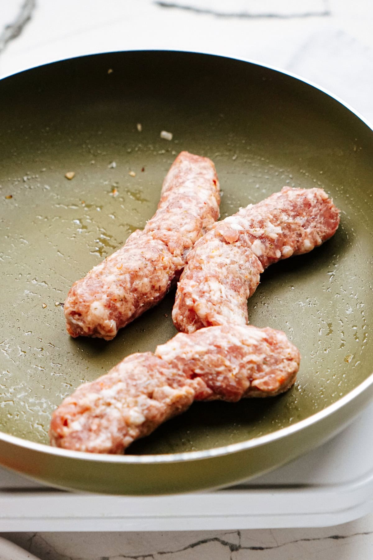 Three uncooked sausages are in a green non-stick frying pan. The sausages are slightly browned on the sides.