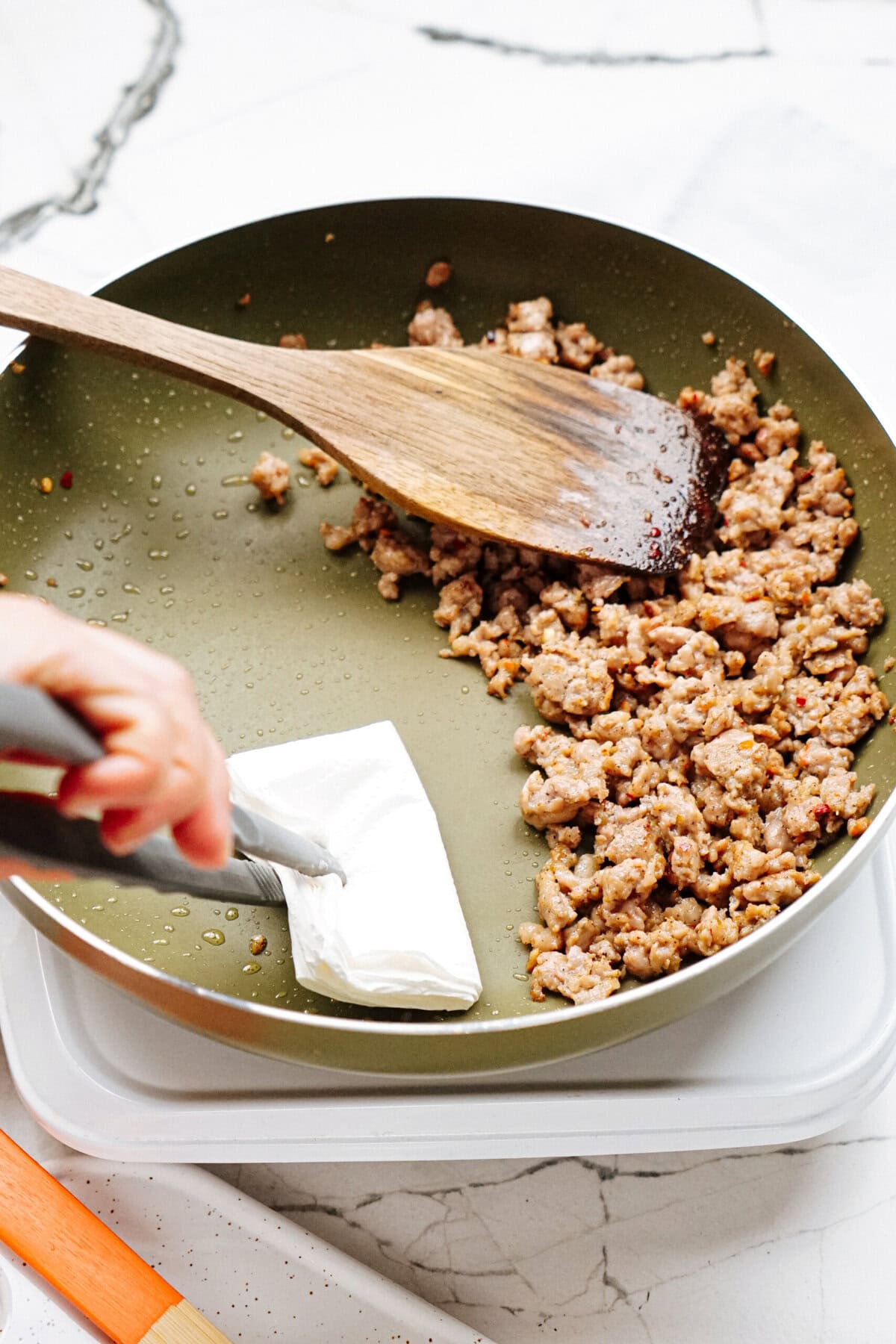 A skillet containing cooked ground sausage, with a wooden spatula and a hand using tongs to hold a block of cream cheese, on a white countertop.