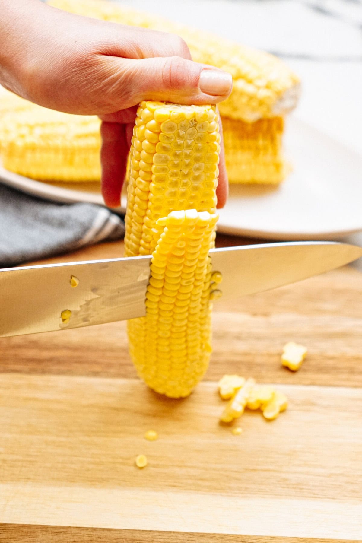 A hand holds a corn cob upright on a wooden surface while a knife slices down to remove kernels. Several corn cobs lie on a plate in the background.