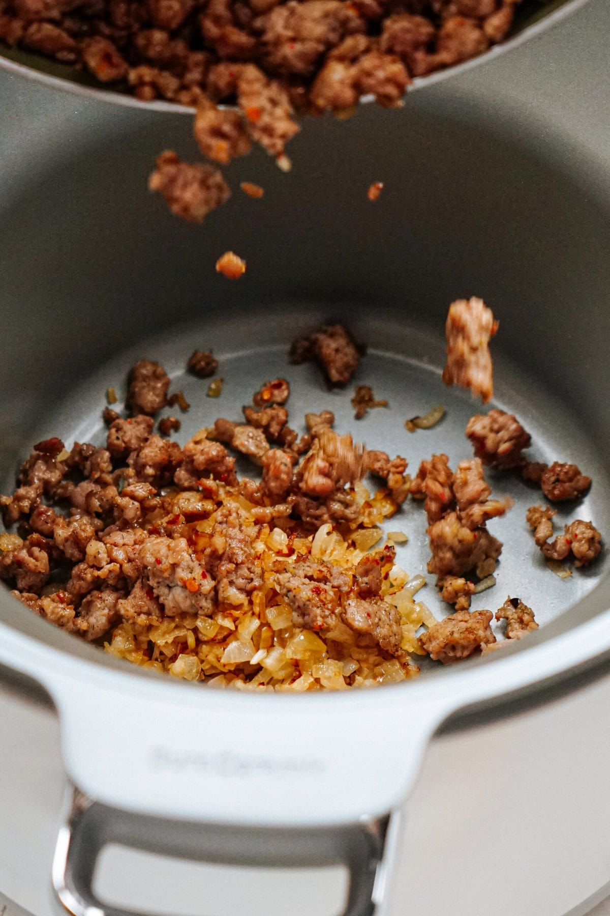 Ground meat and diced onions are sautéing in a white cooking pot. Some of the meat is being added from a skillet above.