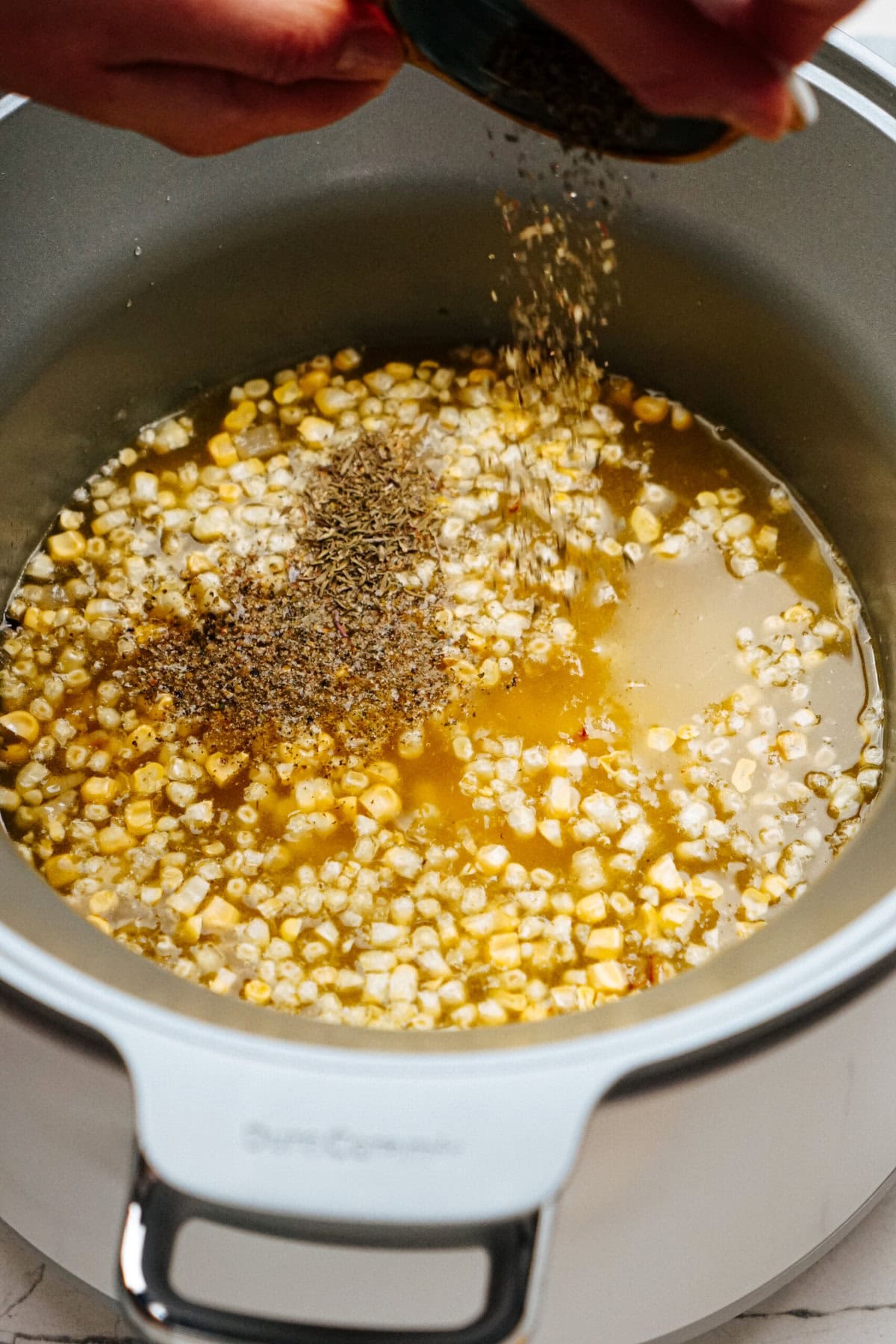 A person sprinkles herbs and spices into a pot of corn and broth.