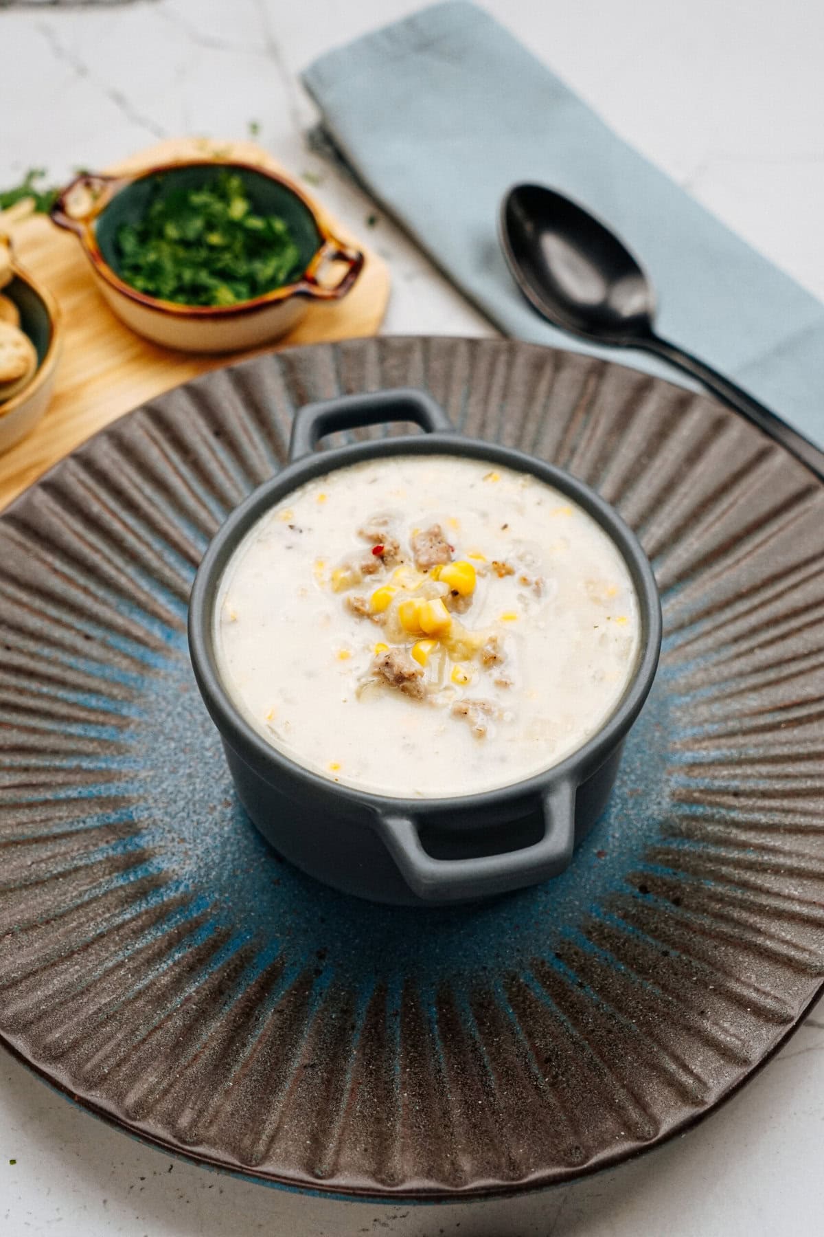 A blue dish filled with a creamy soup containing corn and pieces of meat is placed on a textured gray plate, with a black spoon and a bowl of chopped herbs on the side.